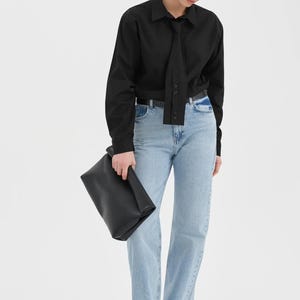 Puede incluir: Una camisa negra de manga larga con detalle de corbata, combinada con vaqueros azul claro y un bolso de mano negro. El conjunto se completa con gafas de sol y zapatos negros. La camisa tiene una botonadura.
