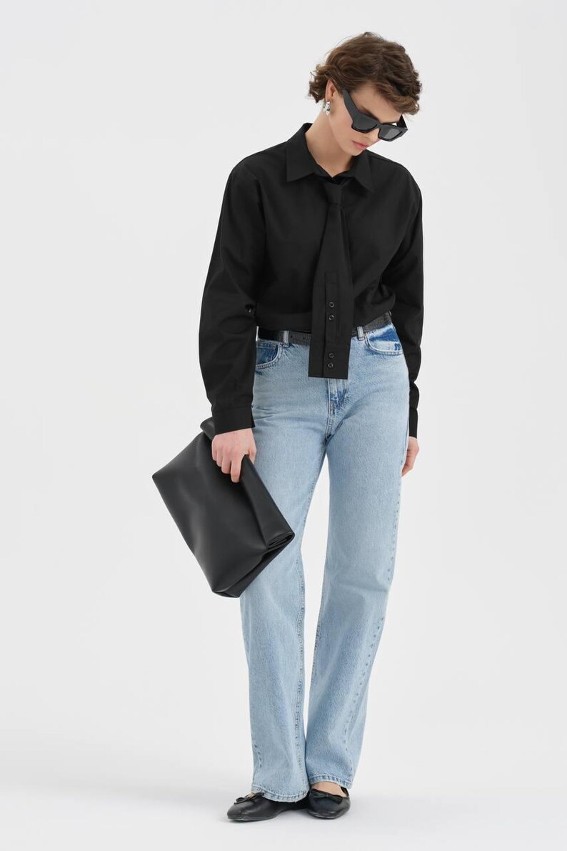 Puede incluir: Una camisa negra de manga larga con detalle de corbata, combinada con vaqueros azul claro y un bolso de mano negro. El conjunto se completa con gafas de sol y zapatos negros. La camisa tiene una botonadura.