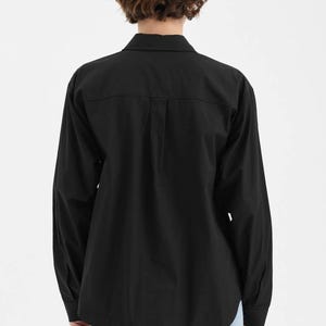 Puede incluir: Una camisa negra de manga larga con botones. La camisa tiene un cuello cl&aacute;sico, pu&ntilde;os abotonados y un dobladillo curvo. El modelo lleva vaqueros azul claro. La camisa est&aacute; hecha de una tela lisa y opaca.