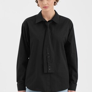 Puede incluir: Camisa negra de manga larga con un dise&ntilde;o &uacute;nico tipo corbata. La camisa presenta un cuello cl&aacute;sico y pu&ntilde;os abotonados. El detalle de la corbata se sujeta con botones. La modelo lleva vaqueros azules.