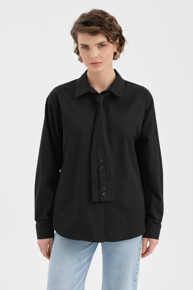 Puede incluir: Camisa negra de manga larga con un dise&ntilde;o &uacute;nico tipo corbata. La camisa presenta un cuello cl&aacute;sico y pu&ntilde;os abotonados. El detalle de la corbata se sujeta con botones. La modelo lleva vaqueros azules.