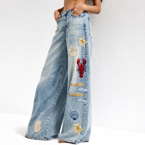 Pode incluir: Jeans azul claro de perna larga com patches bordados com tema marinho. Os patches incluem uma lagosta vermelha, estrelas do mar, conchas e peixes. Os jeans têm uma lavagem desbotada e um corte descontraído.