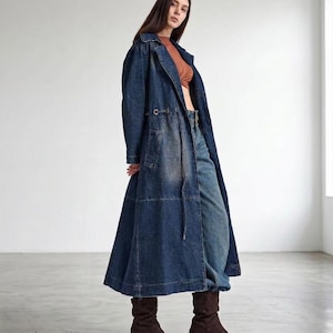 May include: A long, dark blue denim trench coat featuring a collared neckline, long sleeves, and a drawstring waist with metal eyelets. The coat has a structured silhouette, visible seam detailing, and a full-length skirt that falls below the knees.