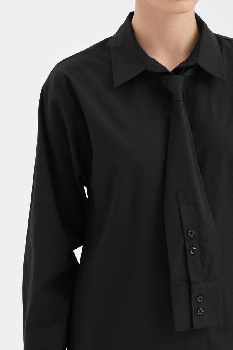 Puede incluir: Camisa negra de manga larga con un detalle de corbata &uacute;nico. La camisa presenta un cuello cl&aacute;sico y pu&ntilde;os abotonados. La corbata est&aacute; integrada en el dise&ntilde;o, creando un look moderno y elegante. La camisa est&aacute; hecha de una tela negra lisa y suave.