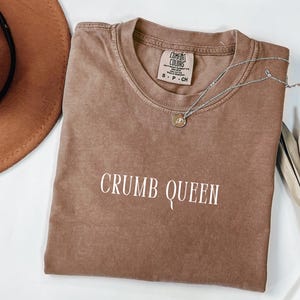 May include: A brown Comfort Colors t-shirt with the text "CRUMB QUEEN" in white. A silver necklace with a small pendant rests on the shirt. A brown hat is partially visible in the upper left corner.