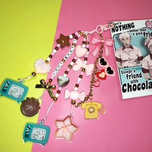 May include: A collection of charms and beads with a pink and gold color scheme. Charms include a TV, a telephone, hearts, bows, and a star. The phrase "I LOVE LUCY" is spelled out in beads. A card with the text "There's NOTHING better than a good friend. Except a friend with Chocolate!" is also included.