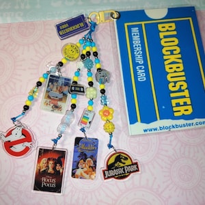 May include: A Blockbuster membership card and a keychain with movie-themed charms. The keychain includes charms of a Ghostbusters logo, a Jurassic Park logo, and movie covers. The keychain has blue, yellow, and black beads.