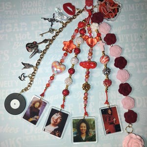 May include: A handmade keychain with multiple charms. It features a red clasp, a vinyl record, photos, beads, and decorative charms like a guitar, lips, and roses. The color palette includes red, pink, gold, and clear accents.