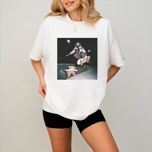 May include: White t-shirt with a square graphic showing a skater and a small green alien on skateboards. The skater wears a helmet and the alien a tan coat. The t-shirt is a casual style, suitable for everyday wear.