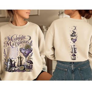 May include: A beige crewneck sweatshirt featuring a graphic print of a margarita glass, a teapot, a frog in a witch hat, and mushrooms. The front displays the text "Midnight Margaritas" with a poem, while the back shows a vertical arrangement of icons.