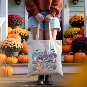 May include: A beige canvas tote bag featuring a colorful graphic print of two blue and pink alien characters dressed in Halloween costumes, including a pumpkin suit and mummy wrappings, set against a backdrop of a castle, bats, and ghosts.