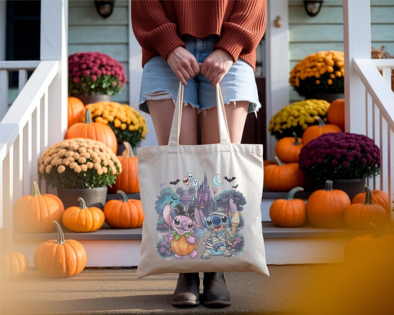 May include: A beige canvas tote bag featuring a colorful graphic print of two blue and pink alien characters dressed in Halloween costumes, including a pumpkin suit and mummy wrappings, set against a backdrop of a castle, bats, and ghosts.