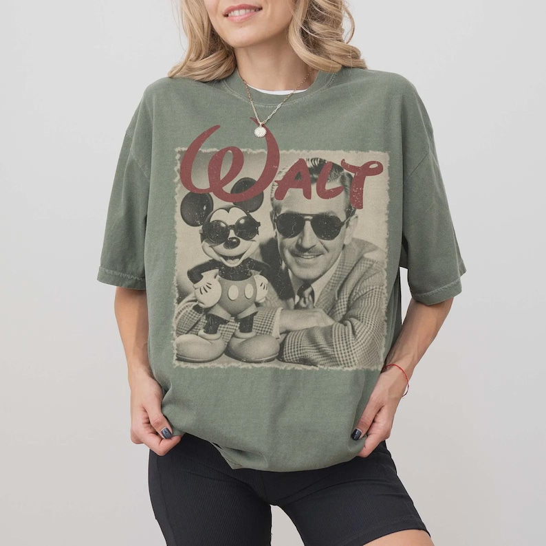 Puede incluir: Una camiseta de manga corta color verde salvia con un estampado gr&aacute;fico de Mickey Mouse y Walt Disney, ambos con gafas de sol. El texto "WALT" aparece en una fuente roja grande y desgastada sobre la imagen fotogr&aacute;fica en blanco y negro.