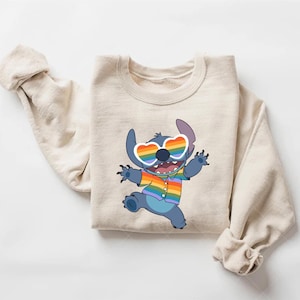 May include: Beige sweatshirt featuring a cartoon character with blue fur, wearing heart-shaped rainbow sunglasses and a rainbow shirt. The character is smiling and has its arms raised. The sweatshirt has long sleeves and a crew neck.