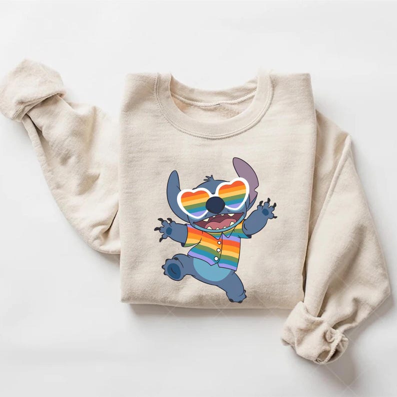 May include: Beige sweatshirt featuring a cartoon character with blue fur, wearing heart-shaped rainbow sunglasses and a rainbow shirt. The character is smiling and has its arms raised. The sweatshirt has long sleeves and a crew neck.