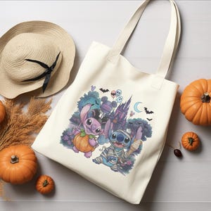 May include: A cream-colored canvas tote bag featuring a colorful graphic of two blue alien characters dressed in Halloween costumes, one as a pumpkin and one as a mummy, set against a backdrop of a castle, bats, and a crescent moon.