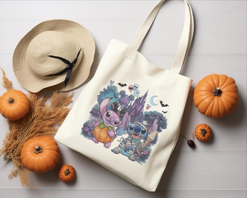 May include: A cream-colored canvas tote bag featuring a colorful graphic of two blue alien characters dressed in Halloween costumes, one as a pumpkin and one as a mummy, set against a backdrop of a castle, bats, and a crescent moon.