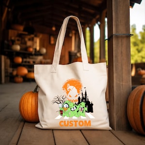 Puede incluir: Un bolso de tela color crema con un estampado gr&aacute;fico de tres personajes de dibujos animados vestidos con disfraces de fantasmas verdes, una silueta negra de un castillo, un &aacute;rbol desnudo, una luna naranja y la palabra CUSTOM en naranja.