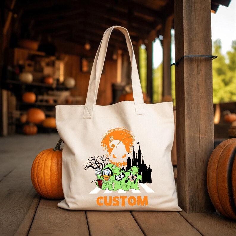 Puede incluir: Un bolso de tela color crema con un estampado gr&aacute;fico de tres personajes de dibujos animados vestidos con disfraces de fantasmas verdes, una silueta negra de un castillo, un &aacute;rbol desnudo, una luna naranja y la palabra CUSTOM en naranja.