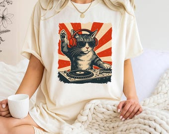 May include: A cream-colored short-sleeve t-shirt featuring a graphic print of a cat wearing red headphones and black sunglasses, positioned behind a DJ turntable. The background of the graphic consists of red and cream-colored sunburst rays.