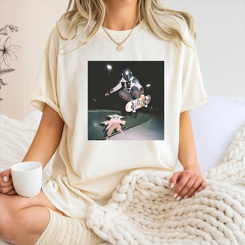 May include: Cream-coloured t-shirt with a graphic of a skater in a helmet performing a trick, and a smaller image of a baby character skateboarding below. The image is set against a dark background.