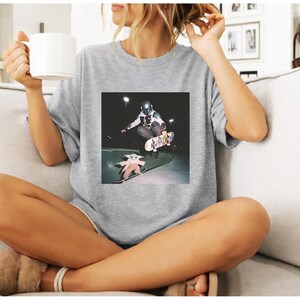 May include: A light grey t-shirt with a square image of a skateboarder doing a trick, with a Baby Yoda figure on a skateboard below. The person in the image is sitting on a sofa and holding a white mug.