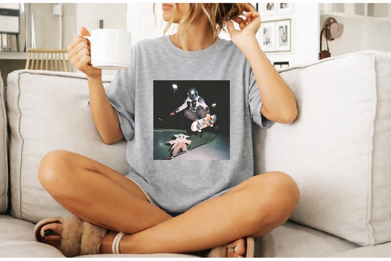 May include: A light grey t-shirt with a square image of a skateboarder doing a trick, with a Baby Yoda figure on a skateboard below. The person in the image is sitting on a sofa and holding a white mug.