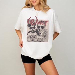 Puede incluir: Una camiseta blanca de manga corta con un estampado gr&aacute;fico de un hombre y un rat&oacute;n de dibujos animados, ambos con gafas de sol. El texto "Walt" est&aacute; impreso en una fuente grande y estilizada de color granate sobre la imagen.