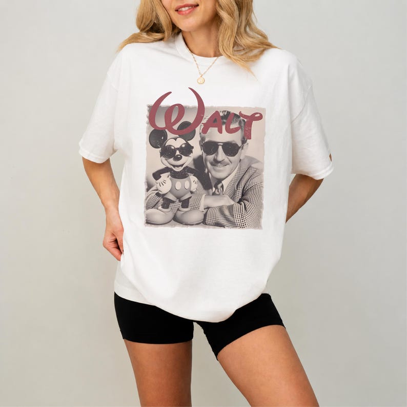 Puede incluir: Una camiseta blanca de manga corta con un estampado gr&aacute;fico de un hombre y un rat&oacute;n de dibujos animados, ambos con gafas de sol. El texto "Walt" est&aacute; impreso en una fuente grande y estilizada de color granate sobre la imagen.