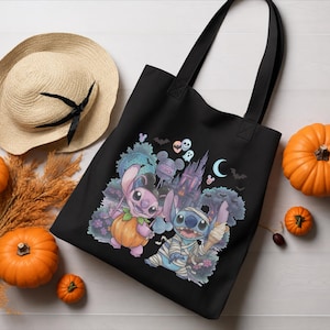 May include: A black tote bag featuring a graphic print of two blue alien characters, one dressed in a pumpkin costume and the other as a mummy, set against a backdrop of a castle, bats, and a crescent moon with the text Studios visible.