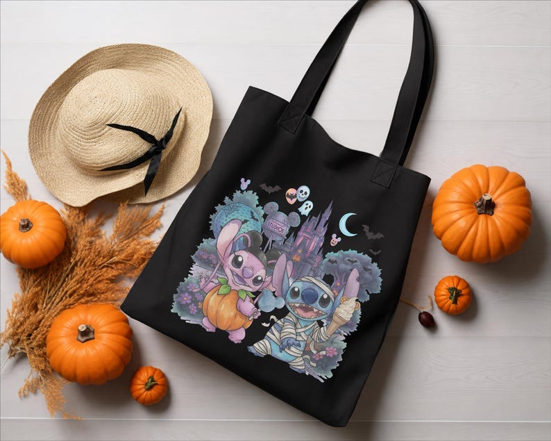 May include: A black tote bag featuring a graphic print of two blue alien characters, one dressed in a pumpkin costume and the other as a mummy, set against a backdrop of a castle, bats, and a crescent moon with the text Studios visible.