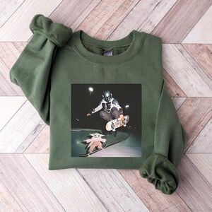 May include: An olive green sweatshirt with a crew neck and long sleeves. The graphic on the front depicts a person in a helmet skateboarding, with a smaller image of a character skateboarding below. The background is a wooden surface.
