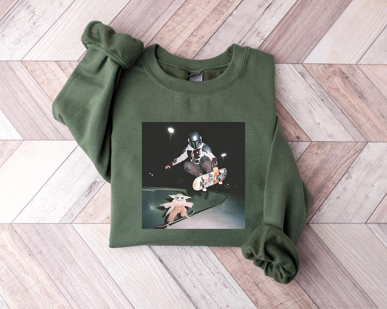 May include: An olive green sweatshirt with a crew neck and long sleeves. The graphic on the front depicts a person in a helmet skateboarding, with a smaller image of a character skateboarding below. The background is a wooden surface.