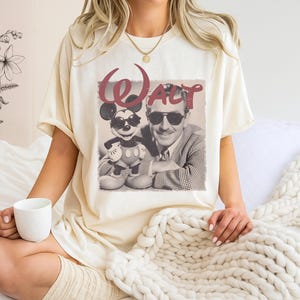 Puede incluir: Una camiseta de manga corta color crema con un estampado gr&aacute;fico de estilo vintage de Walt Disney y Mickey Mouse, ambos con gafas de sol. La palabra Walt est&aacute; impresa en una fuente roja grande y estilizada sobre la imagen en blanco y negro.