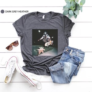 May include: Dark grey heather t-shirt with a graphic of a skateboarder and a small character. The image is set against a dark background. The shirt is paired with blue jeans, white trainers, and sunglasses.
