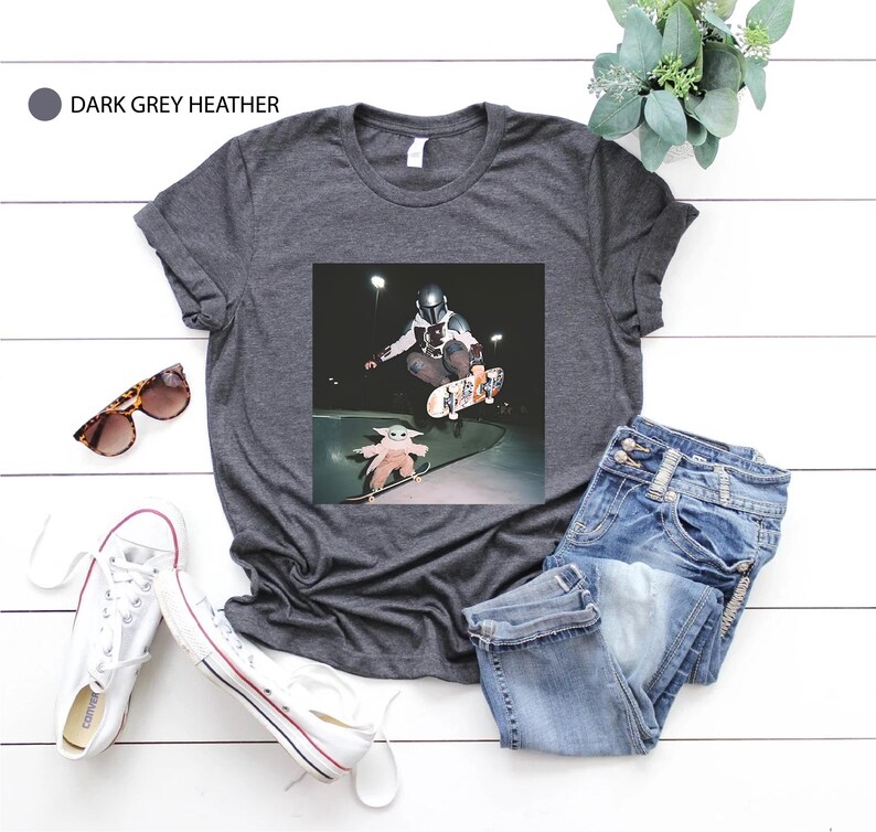 May include: Dark grey heather t-shirt with a graphic of a skateboarder and a small character. The image is set against a dark background. The shirt is paired with blue jeans, white trainers, and sunglasses.