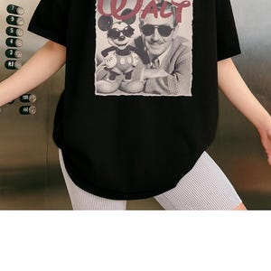 Puede incluir: Una camiseta negra de manga corta con un estampado gr&aacute;fico de Walt Disney y Mickey Mouse, ambos con gafas de sol. El texto "WALT" est&aacute; impreso en una fuente roja estilizada sobre la imagen fotogr&aacute;fica en blanco y negro de las dos figuras.