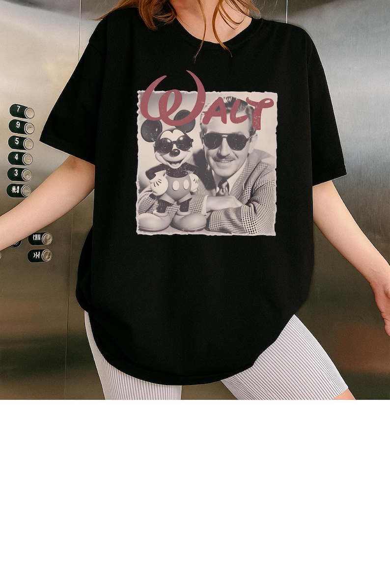 Puede incluir: Una camiseta negra de manga corta con un estampado gr&aacute;fico de Walt Disney y Mickey Mouse, ambos con gafas de sol. El texto "WALT" est&aacute; impreso en una fuente roja estilizada sobre la imagen fotogr&aacute;fica en blanco y negro de las dos figuras.
