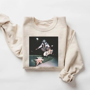 May include: A cream-coloured sweatshirt with a graphic print. The print shows a person in a Mandalorian helmet skateboarding, with a Grogu figure on a skateboard below. The background is dark.