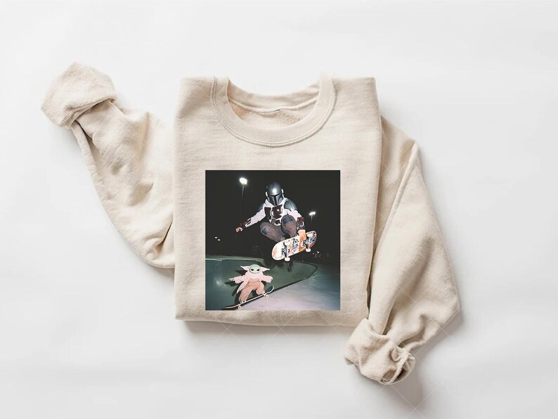 May include: A cream-coloured sweatshirt with a graphic print. The print shows a person in a Mandalorian helmet skateboarding, with a Grogu figure on a skateboard below. The background is dark.