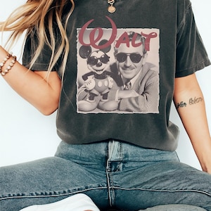 Puede incluir: Una camiseta gris carb&oacute;n de manga corta con un estampado gr&aacute;fico de Mickey Mouse y Walt Disney, ambos con gafas de sol. El texto "WALT" aparece impreso en una fuente roja estilizada sobre las figuras, sobre un fondo cuadrado claro.