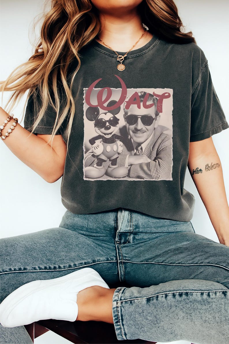 Puede incluir: Una camiseta gris carb&oacute;n de manga corta con un estampado gr&aacute;fico de Mickey Mouse y Walt Disney, ambos con gafas de sol. El texto "WALT" aparece impreso en una fuente roja estilizada sobre las figuras, sobre un fondo cuadrado claro.