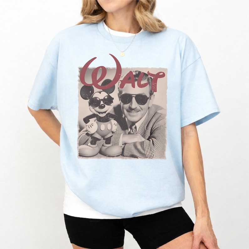 Puede incluir: Una camiseta de manga corta de color azul claro con un estampado gr&aacute;fico en tono sepia de Walt Disney y Mickey Mouse, ambos con gafas de sol. La palabra "WALT" aparece impresa en una fuente grande y estilizada de color granate.