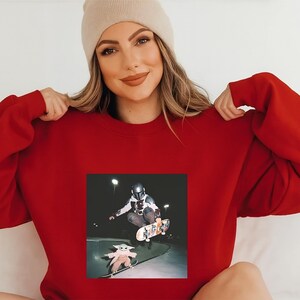 May include: A red sweatshirt featuring a skateboarding scene with a figure in a helmet and a small figure on a skateboard. The small figure is wearing a robe. The person in the image is wearing a beige beanie.