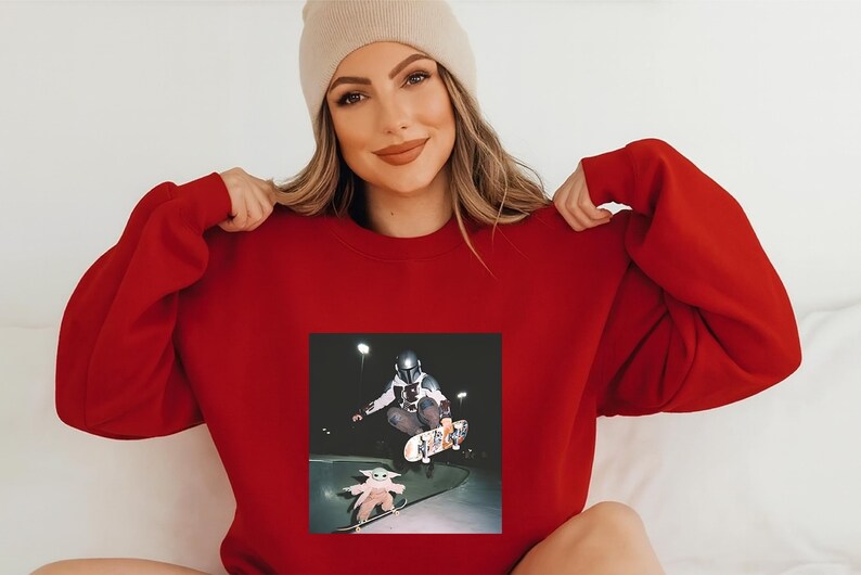 May include: A red sweatshirt featuring a skateboarding scene with a figure in a helmet and a small figure on a skateboard. The small figure is wearing a robe. The person in the image is wearing a beige beanie.