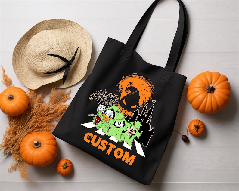 Puede incluir: Un bolso tote negro con un estampado gr&aacute;fico de tres personajes de dibujos animados con disfraces de fantasmas verdes cruzando un paso de peatones. Arriba hay una gran luna naranja con la silueta de un castillo. La palabra CUSTOM est&aacute; en naranja.