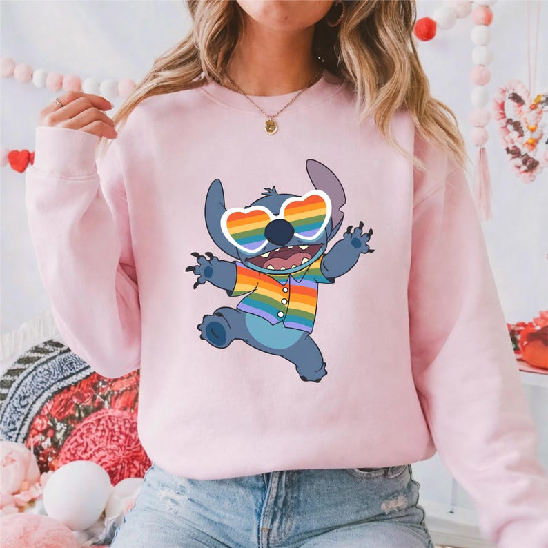 May include: A light pink sweatshirt featuring a cartoon character, Stitch, wearing rainbow heart-shaped sunglasses and a rainbow striped shirt. The character is in a dynamic pose, with arms outstretched and a joyful expression.