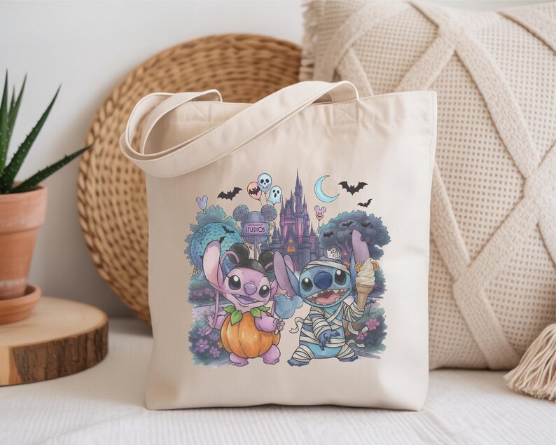 May include: A beige canvas tote bag featuring a colorful graphic print of two blue and pink alien characters dressed in Halloween costumes. One character wears a pumpkin suit, while the other is wrapped in mummy bandages, set against a castle background.