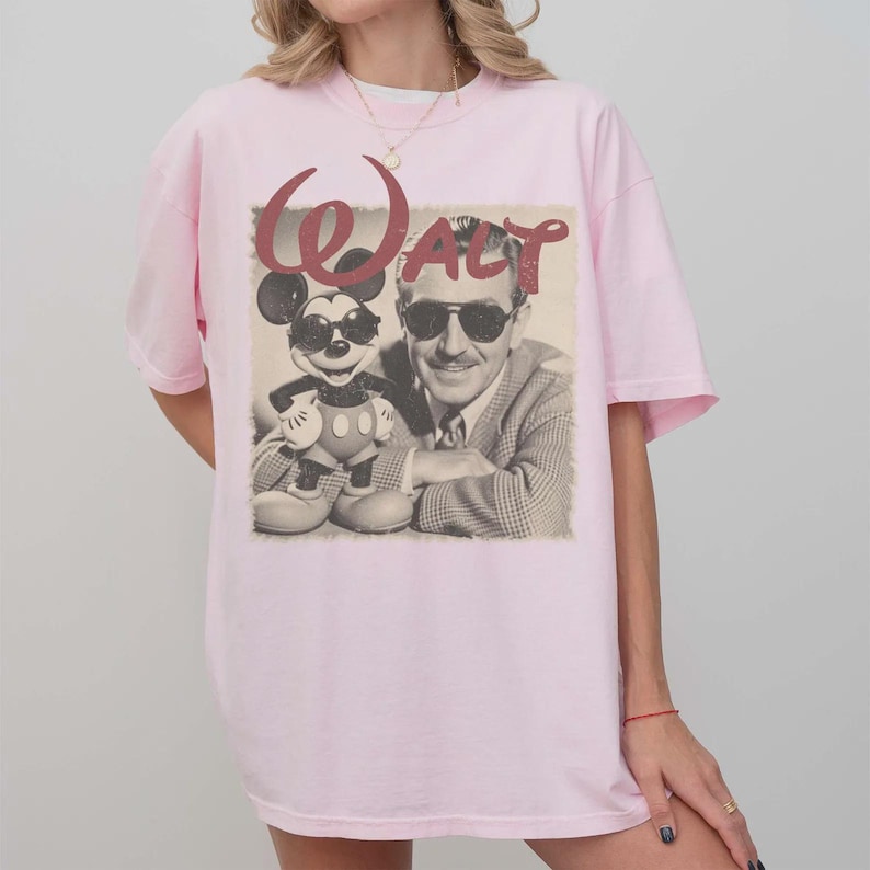 Puede incluir: Una camiseta de manga corta de color rosa claro con un estampado gr&aacute;fico de estilo vintage de Mickey Mouse y Walt Disney, ambos con gafas de sol. El texto Walt aparece en una fuente roja grande y estilizada sobre la imagen en blanco y negro.