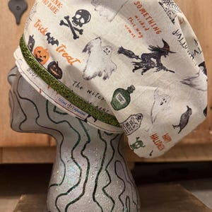 May include: A scrub cap featuring a cream-colored fabric with a Halloween-themed print of ghosts, skulls, pumpkins, witches, and black cats. The cap includes a green glittery band and a small white label with the text Wind Tailored.